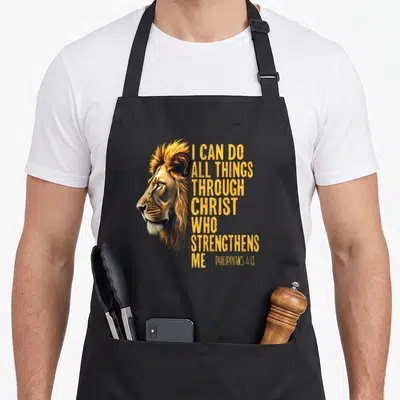 Philippian christian apron - Realistic lion head next to stacked yellow text I CAN DO ALL THINGS THROUGH CHRIST WHO — shown on a black cotton canvas apron against a white background