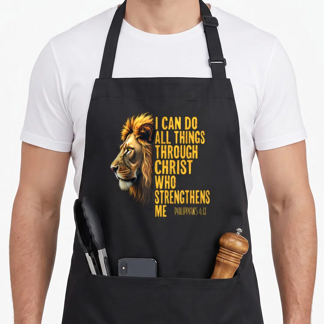 Philippian christian apron - Realistic lion head next to stacked yellow text I CAN DO ALL THINGS THROUGH CHRIST WHO — shown on a black cotton canvas apron against a white background