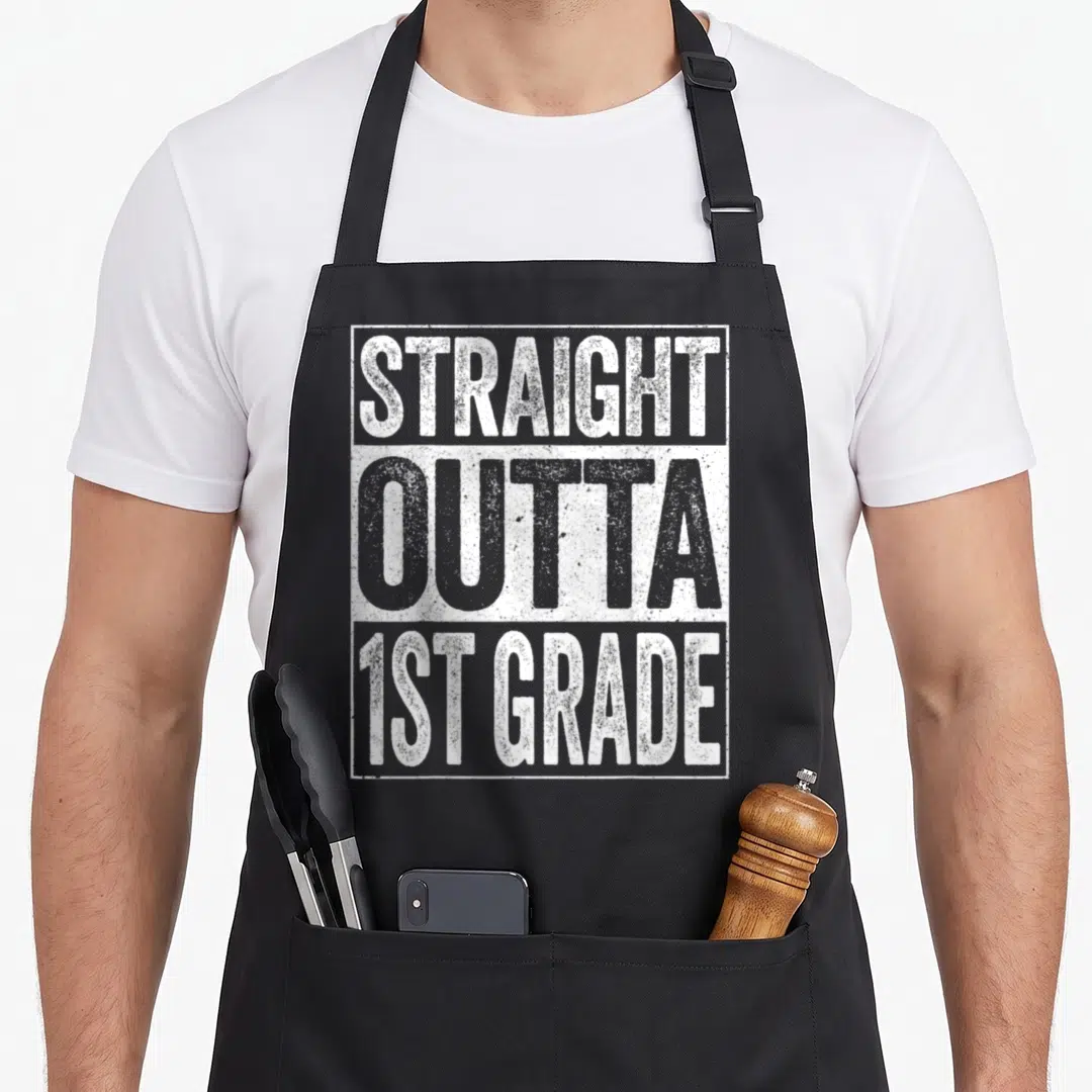 1st straight a student apron - Stacked white and grey distressed text reading STRAIGHT OUTTA 1ST GRADE inside — shown on a black cotton canvas apron against a white background