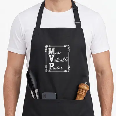 Pastor magnolia apron - White Most Valuable Pastor text in ornate border on black apron — shown on a black cotton canvas apron against a white background