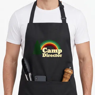 Camper camp chef apron - Beige Camp Director text under a rainbow and dark green pine tree silhouette — shown on a black cotton canvas apron against a white background