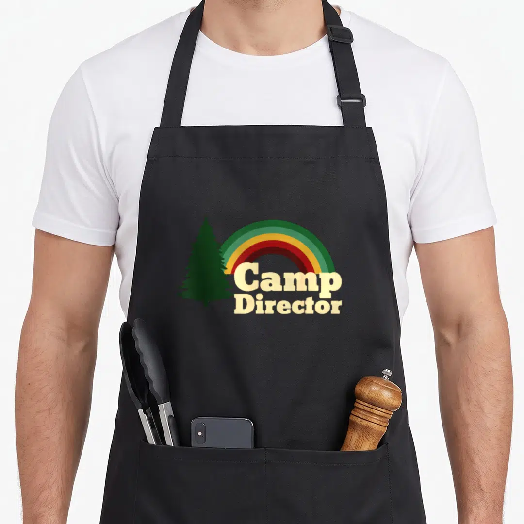 Camper camp chef apron - Beige Camp Director text under a rainbow and dark green pine tree silhouette — shown on a black cotton canvas apron against a white background