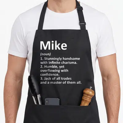 Mike definition birthday apron - White text definition of Mike listing handsome and confident traits in stacked layout — shown on a black cotton canvas apron against a white background