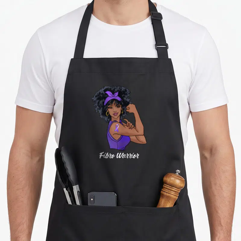Support fibromyalgia awareness apron - Cartoon woman flexing arm with purple ribbon on bicep and white Fibro Warrior — shown on a black cotton canvas apron against a white background
