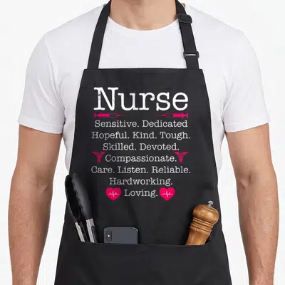 Passion nurse apron - Stacked white text listing nurse virtues flanked by pink syringes, caducei, and heartbeat hearts — shown on a black cotton canvas apron against a white background