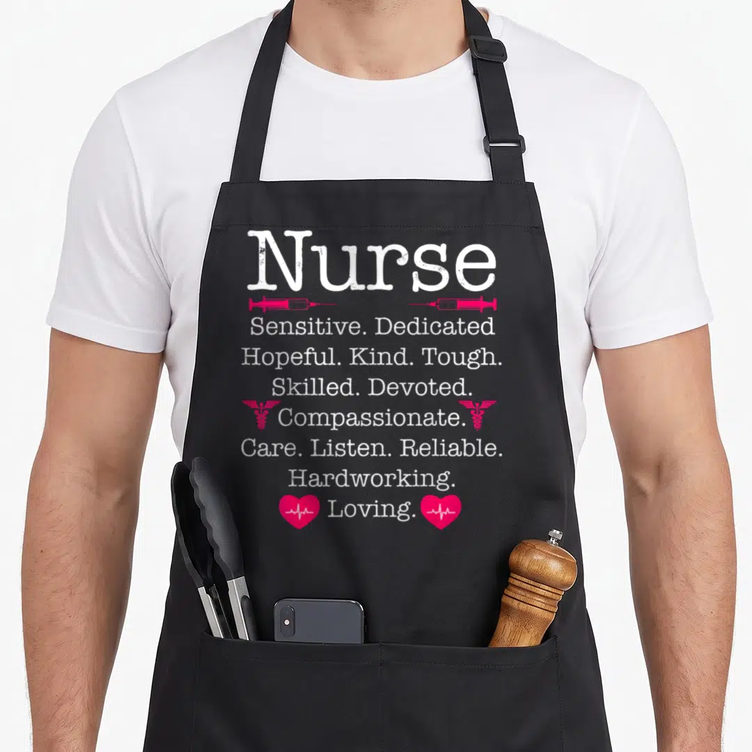 Passion nurse apron - Stacked white text listing nurse virtues flanked by pink syringes, caducei, and heartbeat hearts — shown on a black cotton canvas apron against a white background