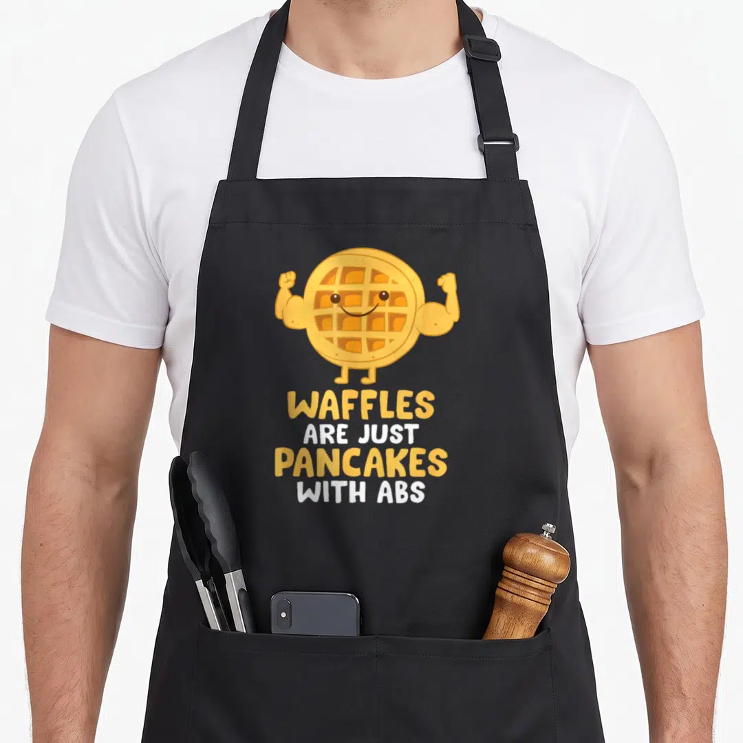 Waffle abs pancake apron - Cartoon waffle flexing muscular arms above yellow and white stacked text reading waffles are — shown on a black cotton canvas apron against a white background