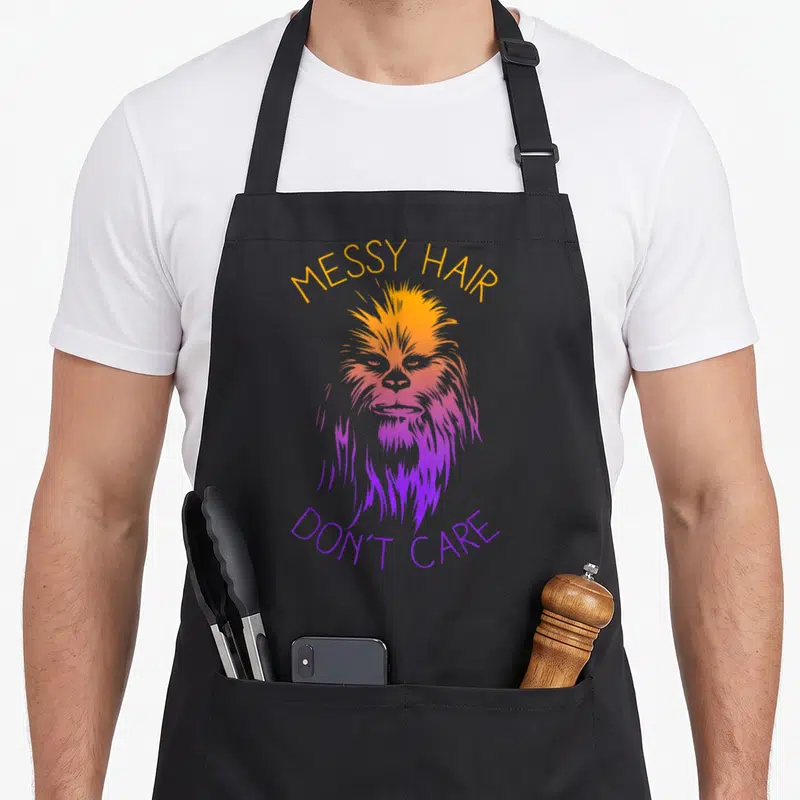 Hair messy aprons - Chewbacca head with MESSY HAIR DON'T CARE text in orange and purple gradient — shown on a black cotton canvas apron against a white background