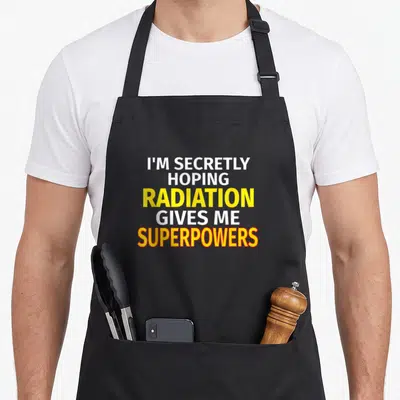 Secretly radiation aprons - Stacked white yellow and orange text reading I'M SECRETLY HOPING RADIATION GIVES ME — shown on a black cotton canvas apron against a white background