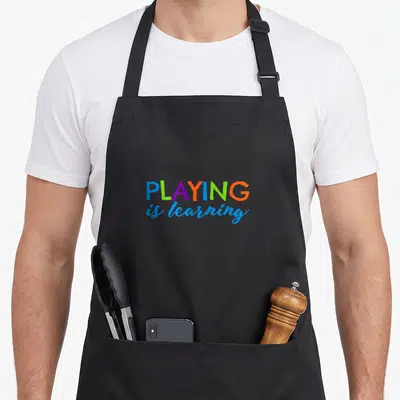 Playing early apron - Stacked colorful text PLAYING is learning in blue green purple red orange typography — shown on a black cotton canvas apron against a white background