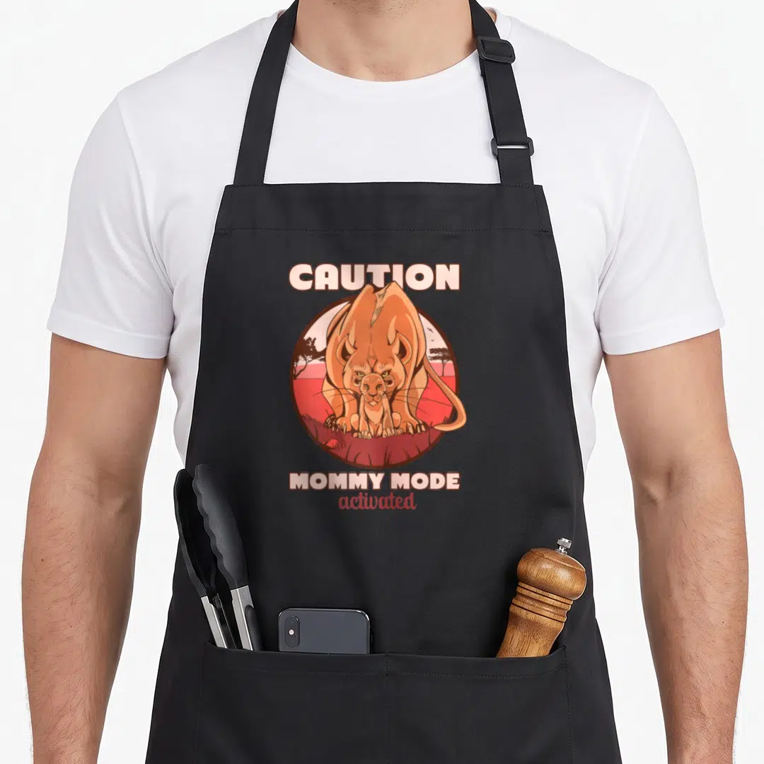 Caution lion apron - Cartoon lioness protecting cub under Caution Mommy Mode activated text — shown on a black cotton canvas apron against a white background