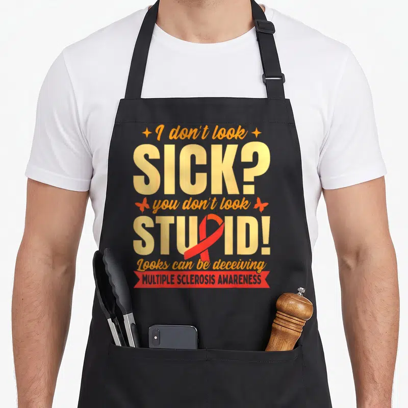 Cute multiple sclerosis awareness apron - Yellow and orange text reading I don't look SICK with red ribbon overlapping — shown on a black cotton canvas apron against a white background