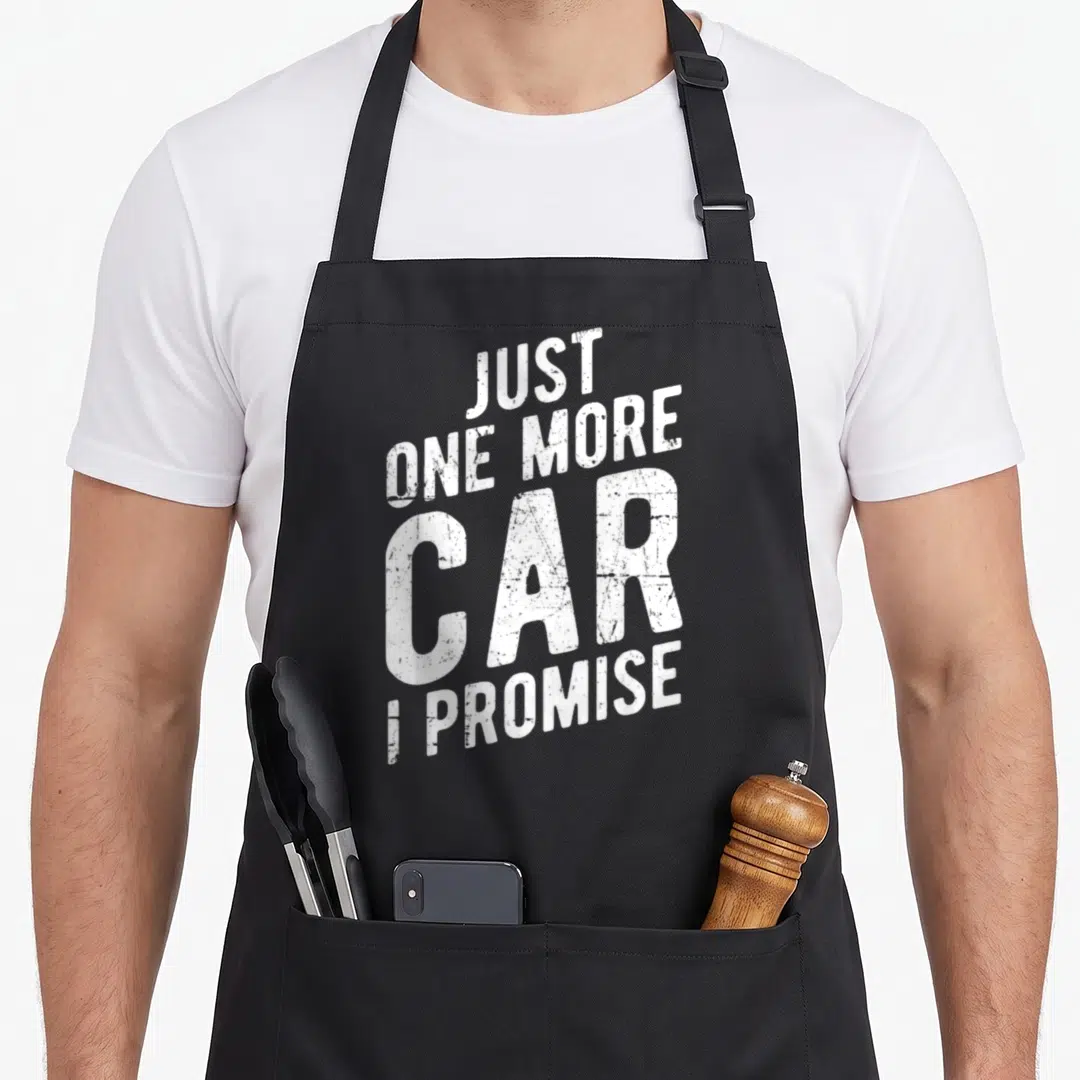 Gear car apron - White distressed text reading Just One More Car I Promise stacked on black fabric — shown on a black cotton canvas apron against a white background