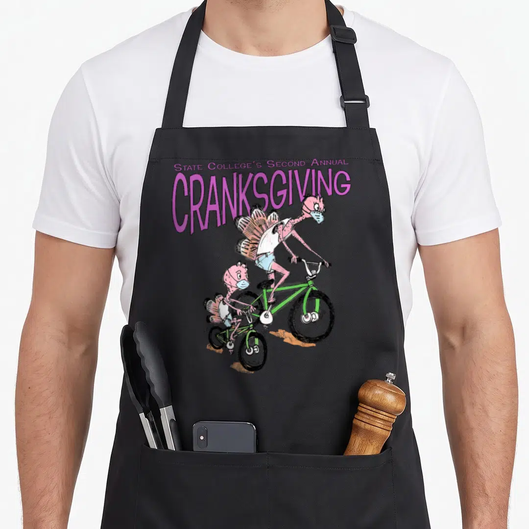 Cranksgiving 2nd apron - Purple text above two pink turkeys riding green bicycles wearing masks — shown on a black cotton canvas apron against a white background