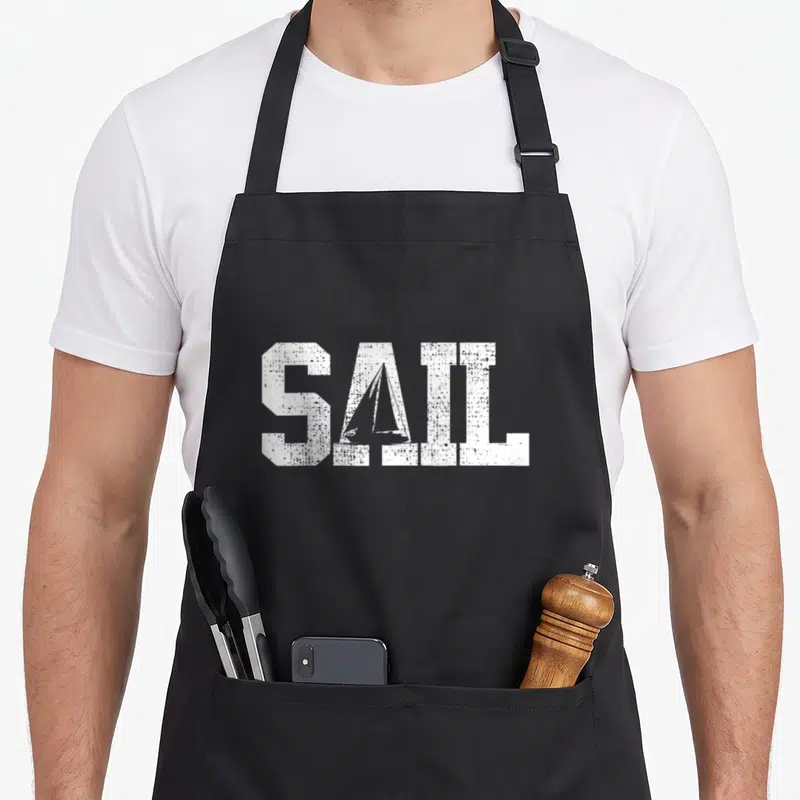 Sail sailing apron - White SAIL text with sailboat silhouette inside the A, distressed texture on black fabric — shown on a black cotton canvas apron against a white background