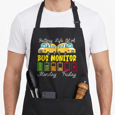 Monitor school bus apron - Two yellow buses above a battery icon draining from green to red plug with text — shown on a black cotton canvas apron against a white background