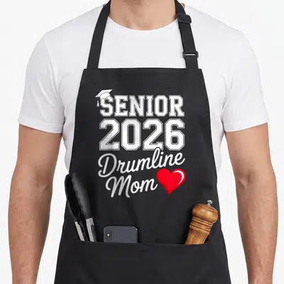 Senior band mom apron - Stacked white text SENIOR 2026 Drumline Mom with graduation cap and red heart — shown on a black cotton canvas apron against a white background