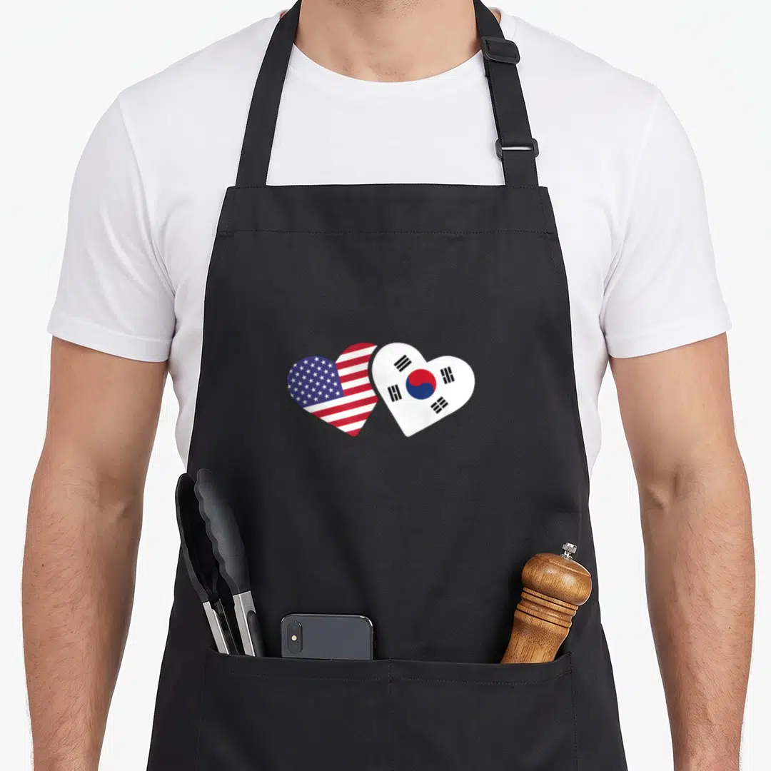 South korea american flag apron - Two heart shapes filled with US and South Korean flag patterns in red white and blue — shown on a black cotton canvas apron against a white background