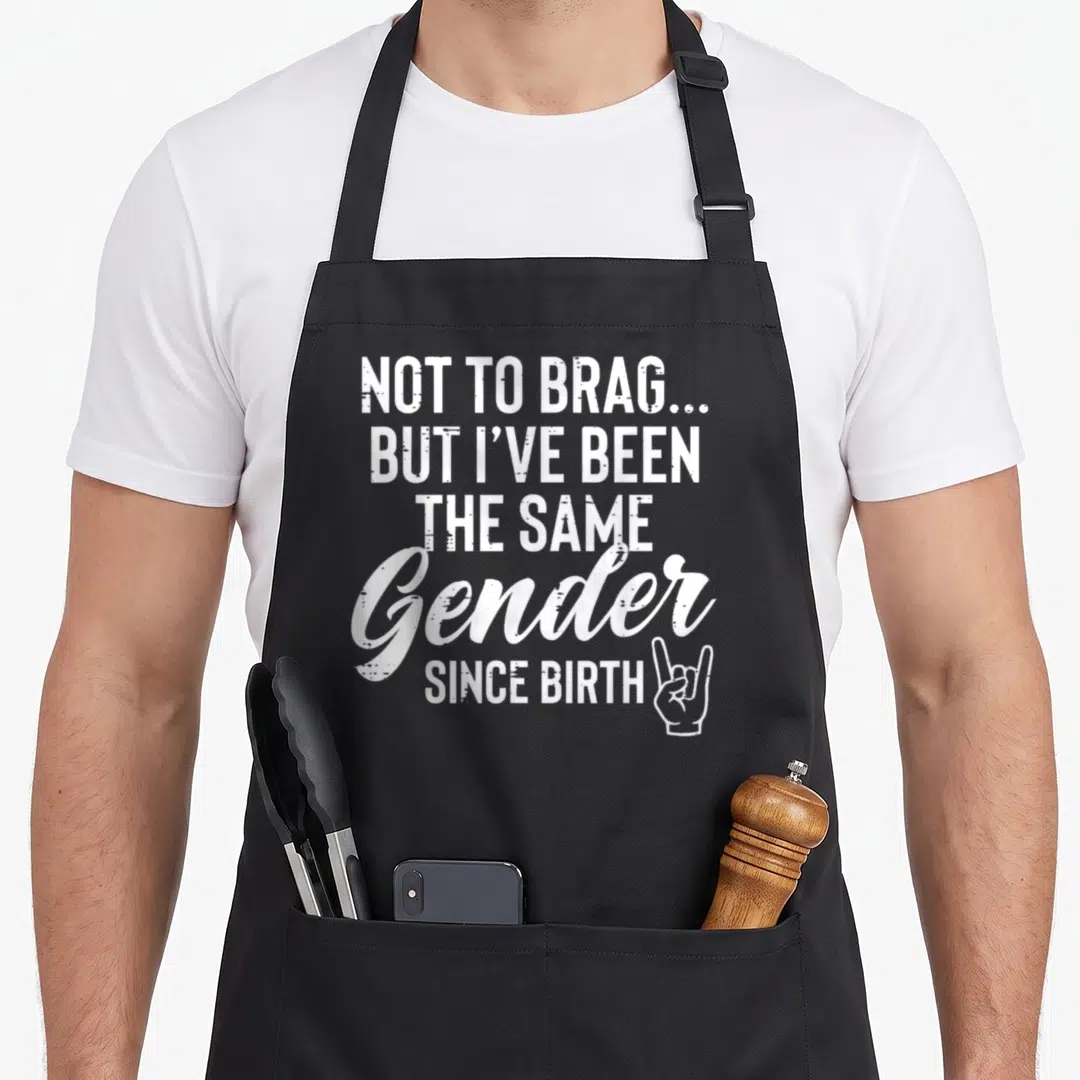 Brag birth apron - White stacked text saying Same Gender Since Birth with rock-on hand symbol — shown on a black cotton canvas apron against a white background