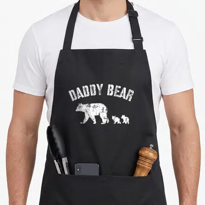 Cub daddy apron - Arched DADDY BEAR text above white bear and two cubs walking left — shown on a black cotton canvas apron against a white background