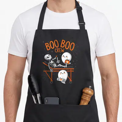 Boo halloween nurse apron - Cartoon ghosts treating a skeleton on an orange gurney with arched BOO BOO CREW text — shown on a black cotton canvas apron against a white background