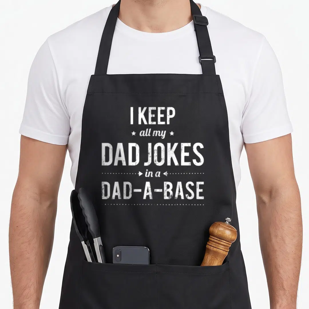 Humorous sarcastic father day apron - White stacked text reading I KEEP all my DAD JOKES in a DAD-A-BASE with stars — shown on a black cotton canvas apron against a white background