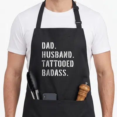 Daughter tattoo aprons - White distressed text reading DAD HUSBAND TATTOOED BADASS stacked vertically — shown on a black cotton canvas apron against a white background