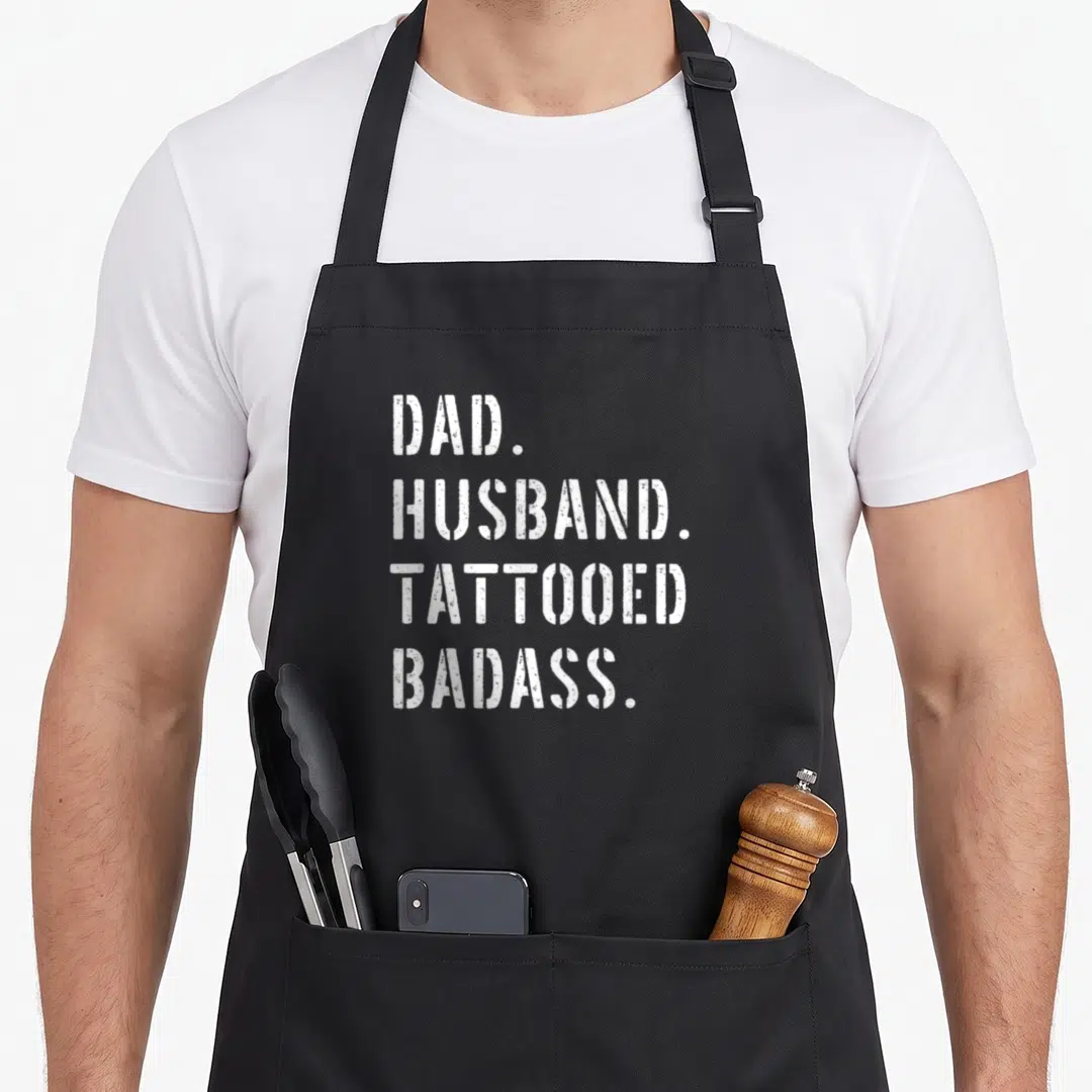 Daughter tattoo aprons - White distressed text reading DAD HUSBAND TATTOOED BADASS stacked vertically — shown on a black cotton canvas apron against a white background