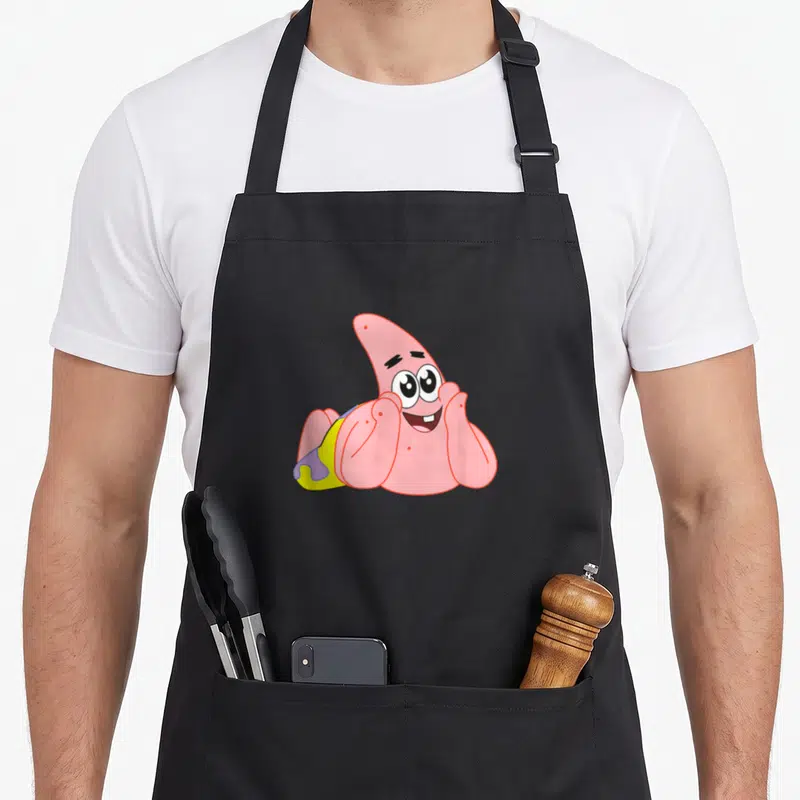 Feelin patrick apron - Pink cartoon starfish lying down with yellow shorts and purple details — shown on a black cotton canvas apron against a white background