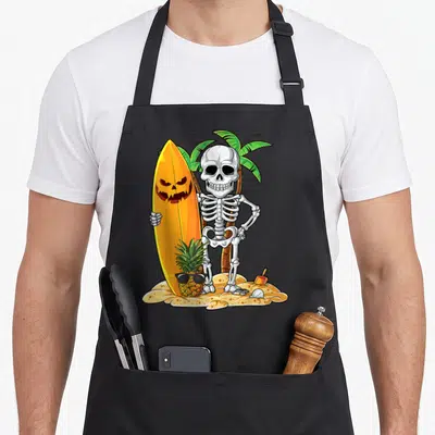 Surfing halloween apron - Cartoon skeleton holding pumpkin surfboard next to sunglass pineapple on tan sand with palm — shown on a black cotton canvas apron against a white background