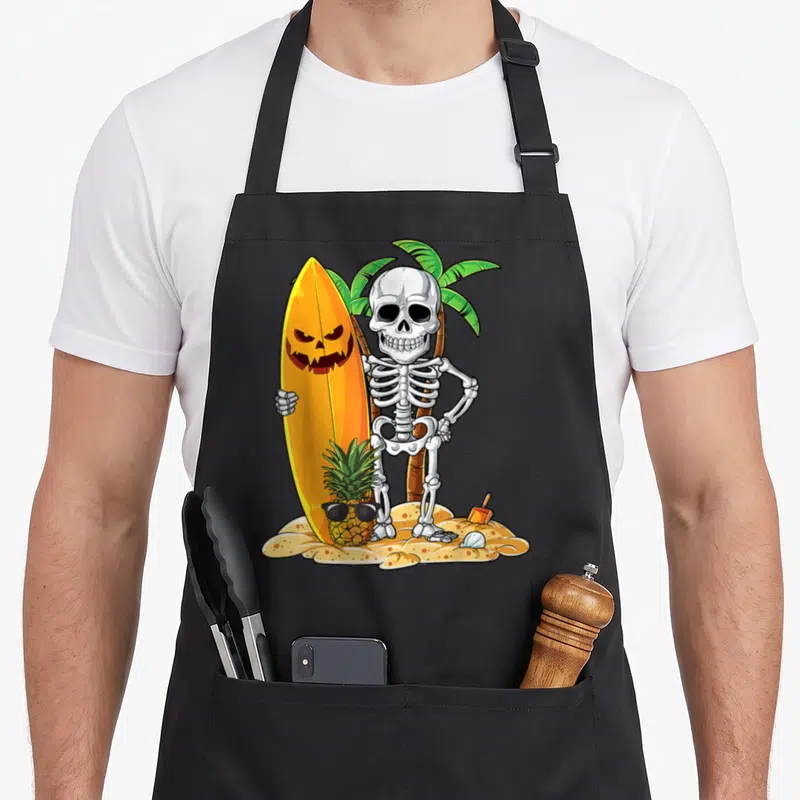 Surfing halloween apron - Cartoon skeleton holding pumpkin surfboard next to sunglass pineapple on tan sand with palm — shown on a black cotton canvas apron against a white background