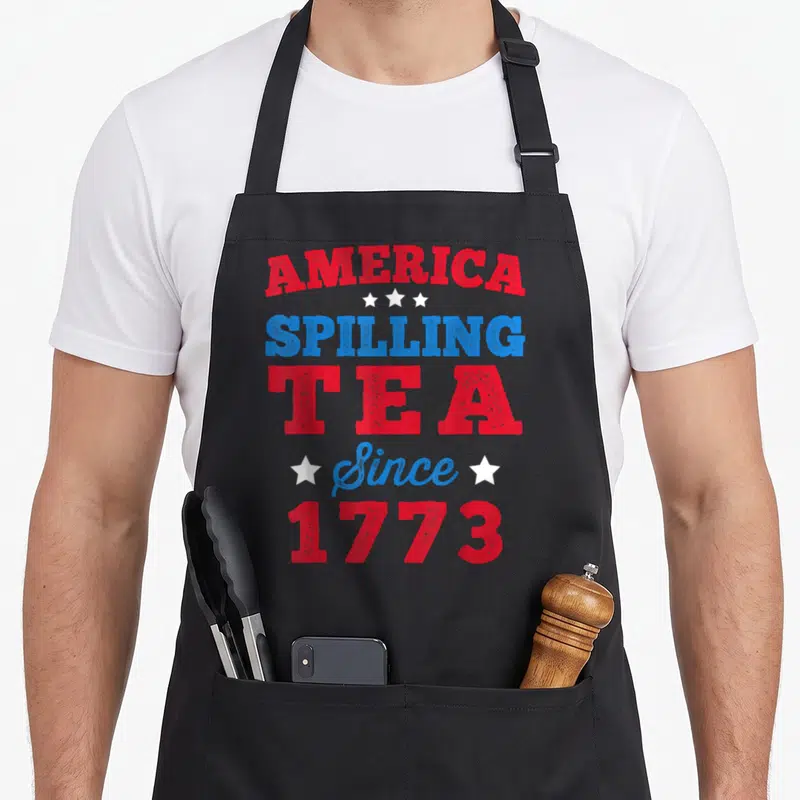 4th tea party aprons - Red and blue stacked text America Spilling Tea Since 1773 with white stars — shown on a black cotton canvas apron against a white background
