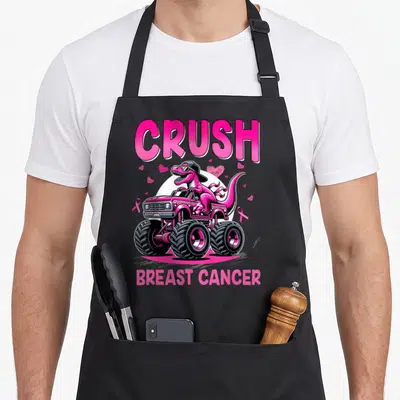 Crush breast cancer awareness aprons - Pink dinosaur wearing hat driving pink monster truck with hearts and ribbons — shown on a black cotton canvas apron against a white background