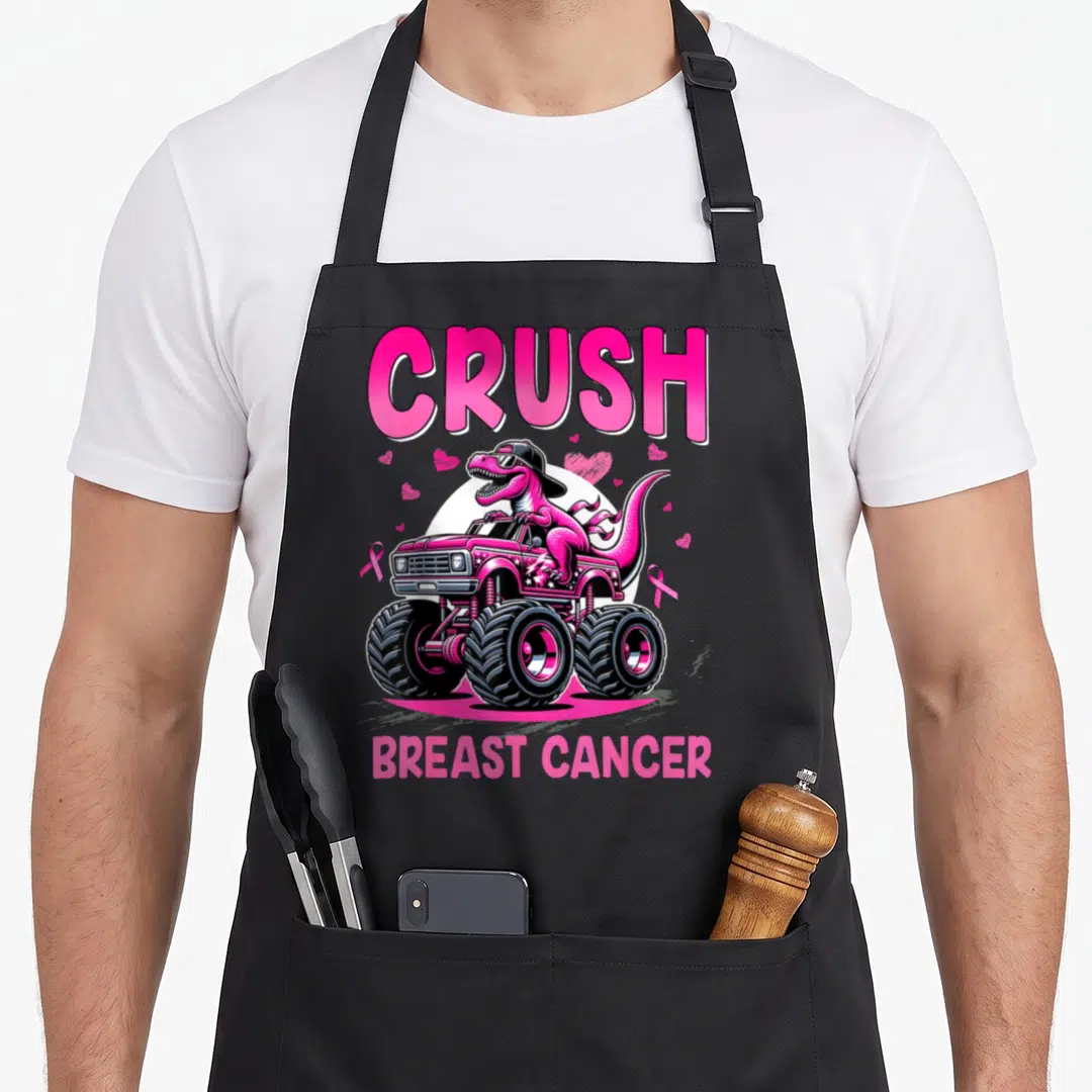 Crush breast cancer awareness aprons - Pink dinosaur wearing hat driving pink monster truck with hearts and ribbons — shown on a black cotton canvas apron against a white background