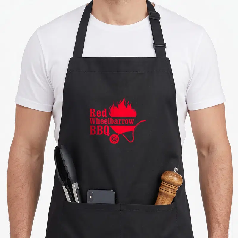 Wheelbarrow mr apron - Red stacked text reading Red Wheelbarrow BBQ beside a cartoon wheelbarrow with flames rising — shown on a black cotton canvas apron against a white background