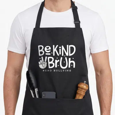 Kind unity day apron - White stacked text Be KiND BrUh with peace sign hand and small hearts on black fabric — shown on a black cotton canvas apron against a white background