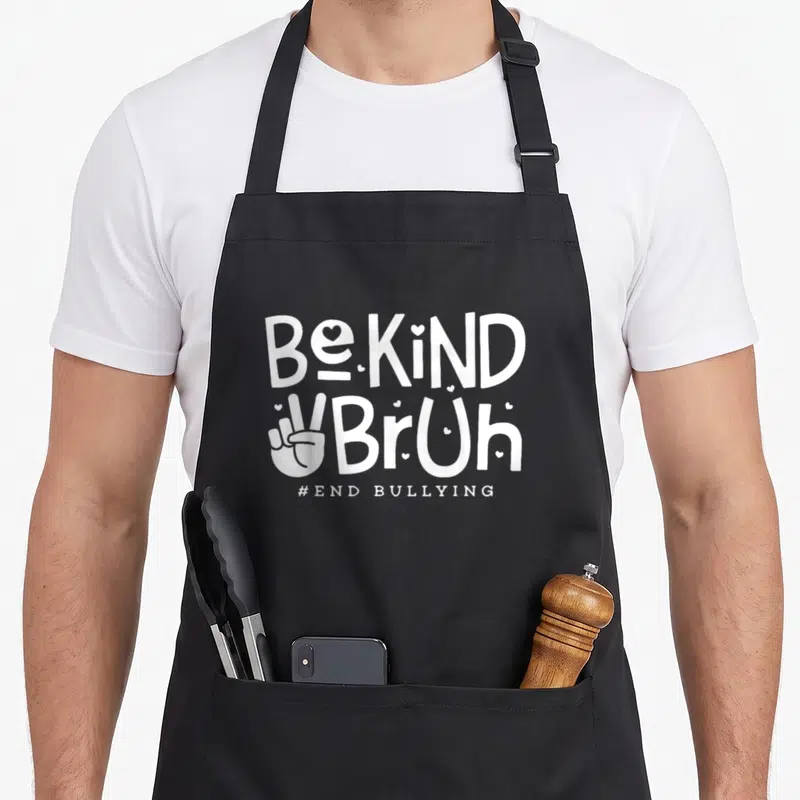 Kind unity day apron - White stacked text Be KiND BrUh with peace sign hand and small hearts on black fabric — shown on a black cotton canvas apron against a white background