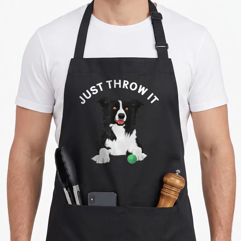 Funny border collie apron - Cartoon black and white dog with green ball and JUST THROW IT text — shown on a black cotton canvas apron against a white background
