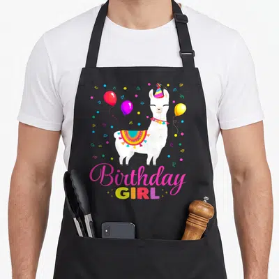 Llama animal birthday apron - White llama wearing party hat and saddle surrounded by balloons and confetti with text — shown on a black cotton canvas apron against a white background