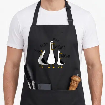 Seagull mother's day aprons - Three cartoon seagulls with open beaks next to MOM text — shown on a black cotton canvas apron against a white background
