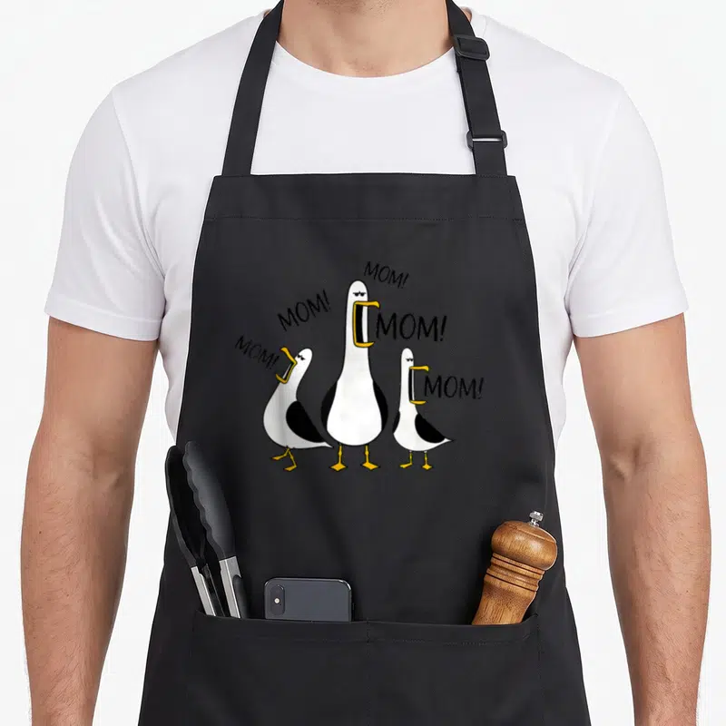 Seagull mother's day aprons - Three cartoon seagulls with open beaks next to MOM text — shown on a black cotton canvas apron against a white background