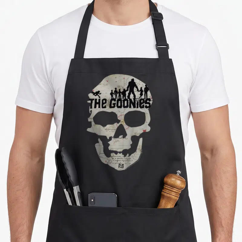 Poster skeleton apron - Skull silhouette filled with map pattern, THE GOONies text, character silhouettes on forehead — shown on a black cotton canvas apron against a white background