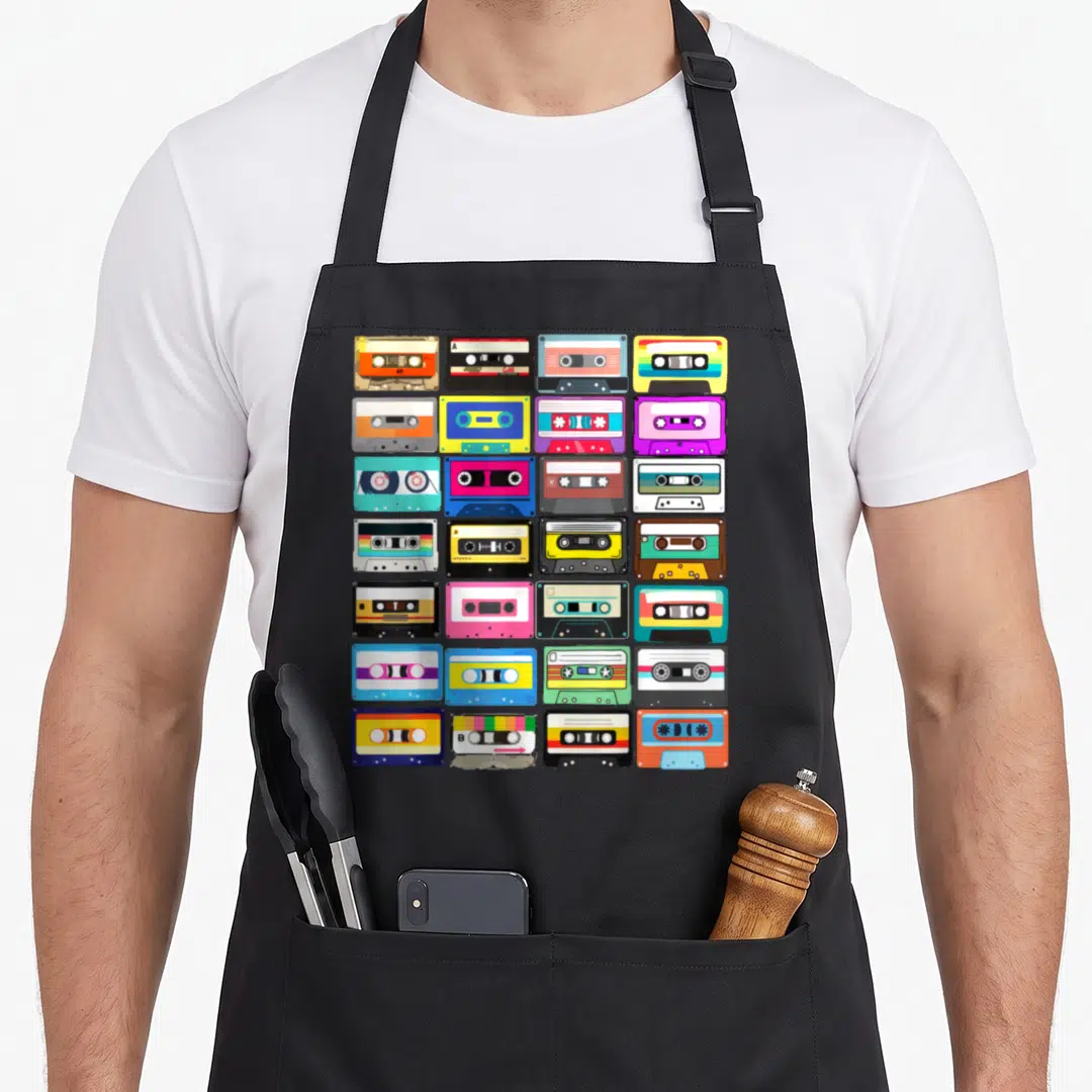 Cassette 1980s apron - Cartoon grid of 24 colorful cassette tapes in blue, yellow, pink, green, and black — shown on a black cotton canvas apron against a white background