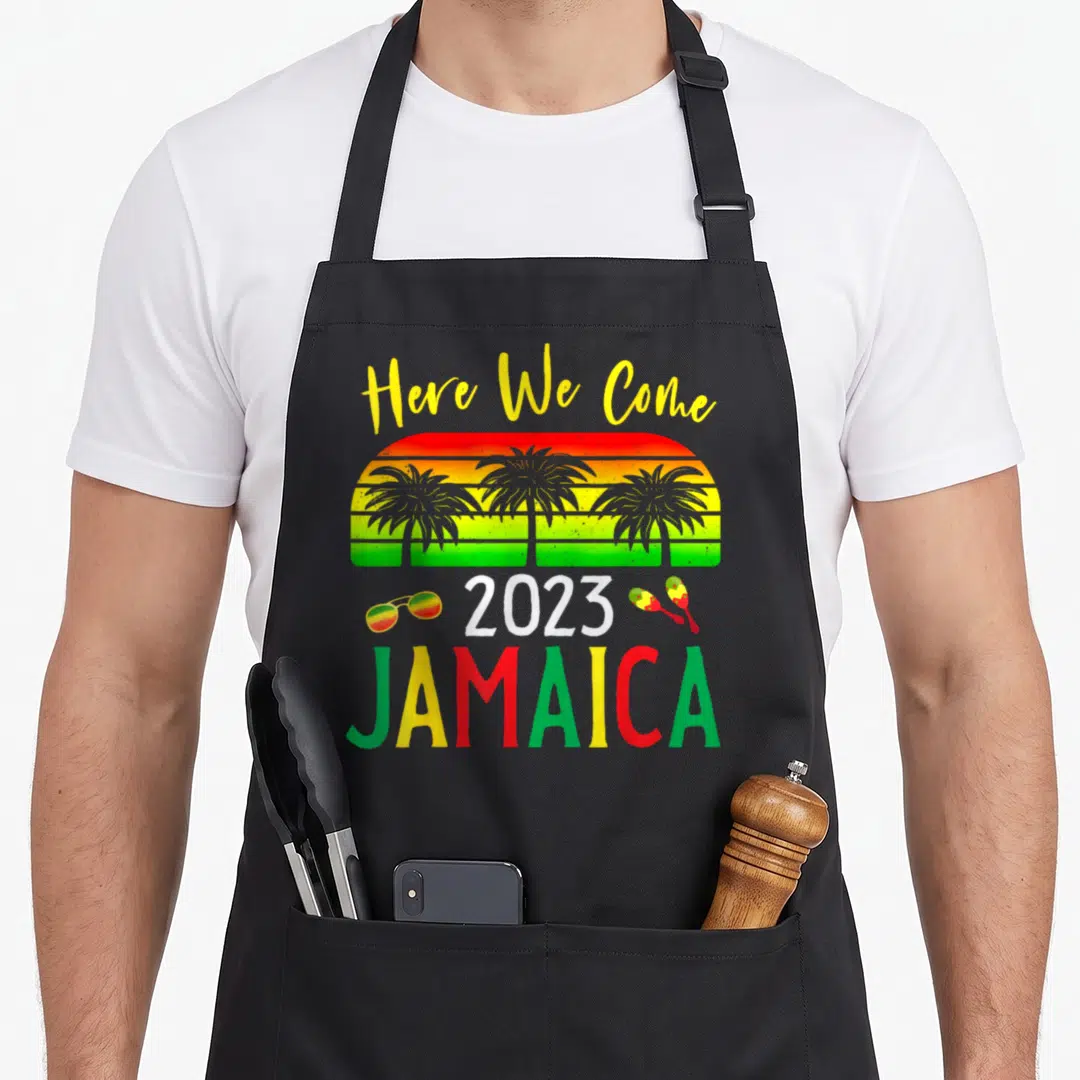 Family come and get it apron - Stacked Here We Come 2023 Jamaica text with palm tree sun, sunglasses, and maracas — shown on a black cotton canvas apron against a white background
