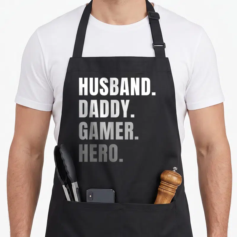 Gamer husband apron - Stacked white and grey text reading HUSBAND. DADDY. GAMER. HERO — shown on a black cotton canvas apron against a white background