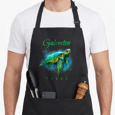 Sea texas apron - Watercolor sea turtle with Galveston and Texas text in green blue and yellow — shown on a black cotton canvas apron against a white background