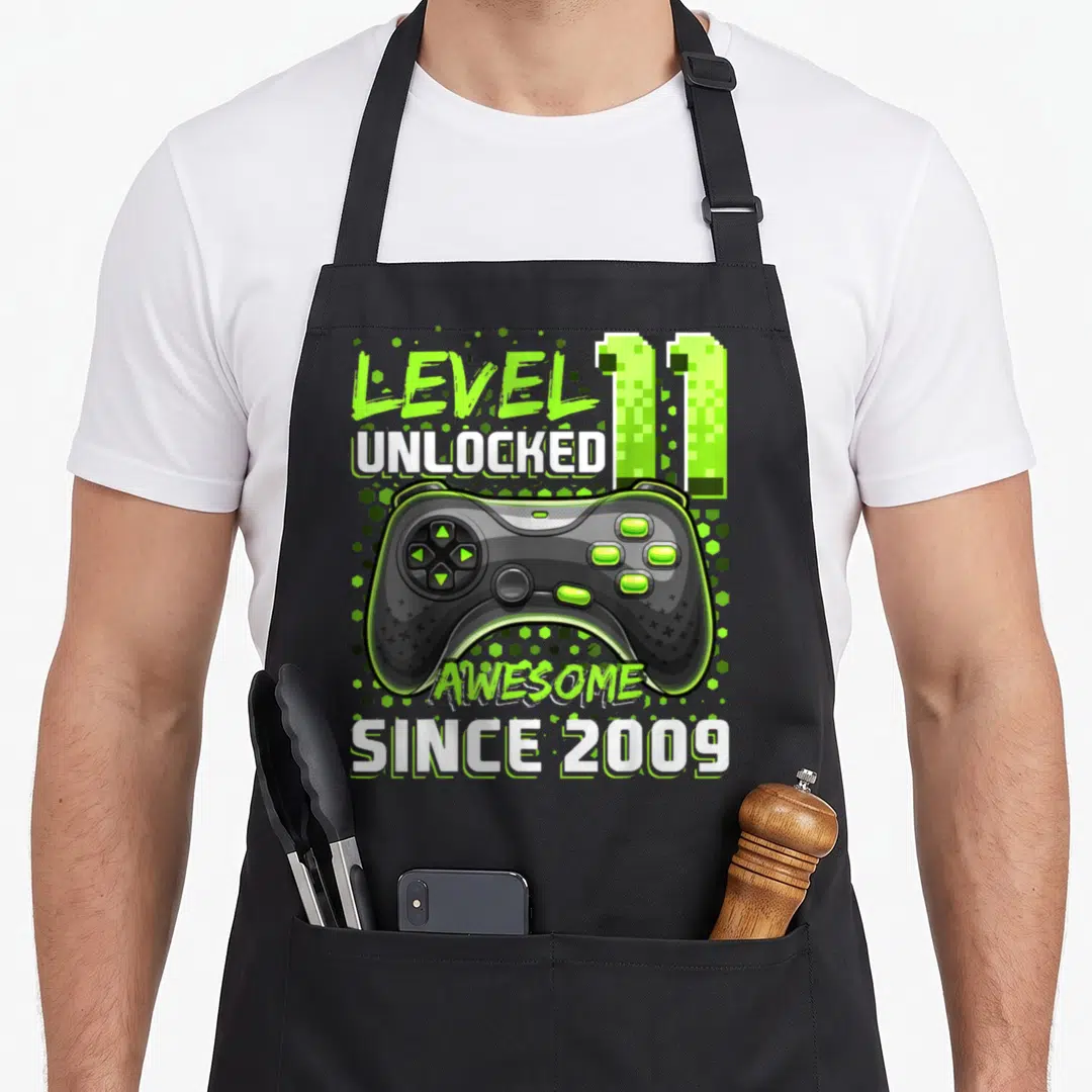 11th birthday age video game apron - Lime green game controller with LEVEL 11 UNLOCKED AWESOME SINCE 2009 text — shown on a black cotton canvas apron against a white background