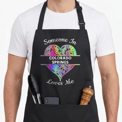 Hometown rainbow apron - Rainbow lace heart with Someone In Colorado Springs Loves Me script text — shown on a black cotton canvas apron against a white background