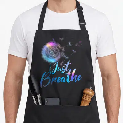 Dandelion breathe apron - Watercolor dandelion with blue purple pink seeds and Just Breathe script — shown on a black cotton canvas apron against a white background