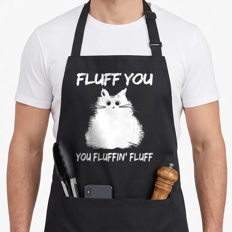 Fluffin cat kitten apron - White cartoon cat between text FLUFF YOU and YOU FLUFFIN' FLUFF — shown on a black cotton canvas apron against a white background